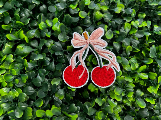 Embroidered patch of two red cherries with a pink bow on a green leafy background