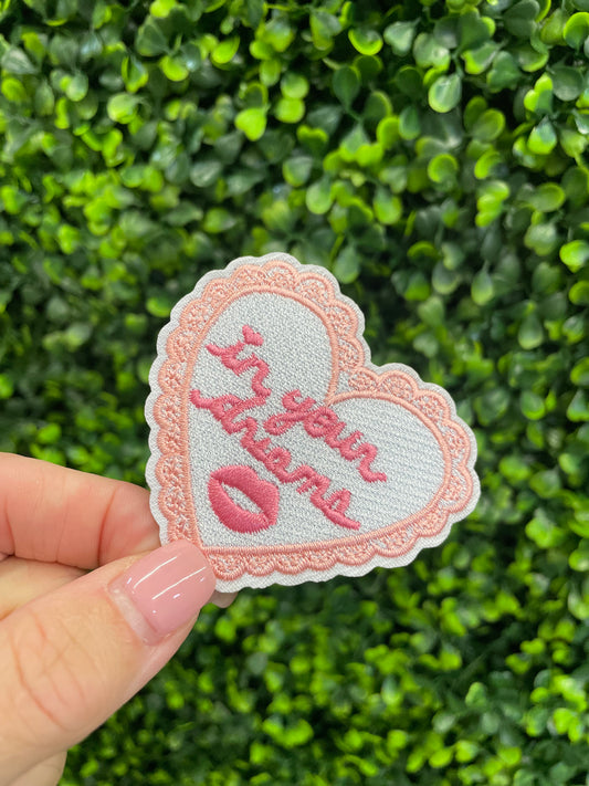 Heart-shaped embroidered patch with 'In your dreams' text held against a green leafy background