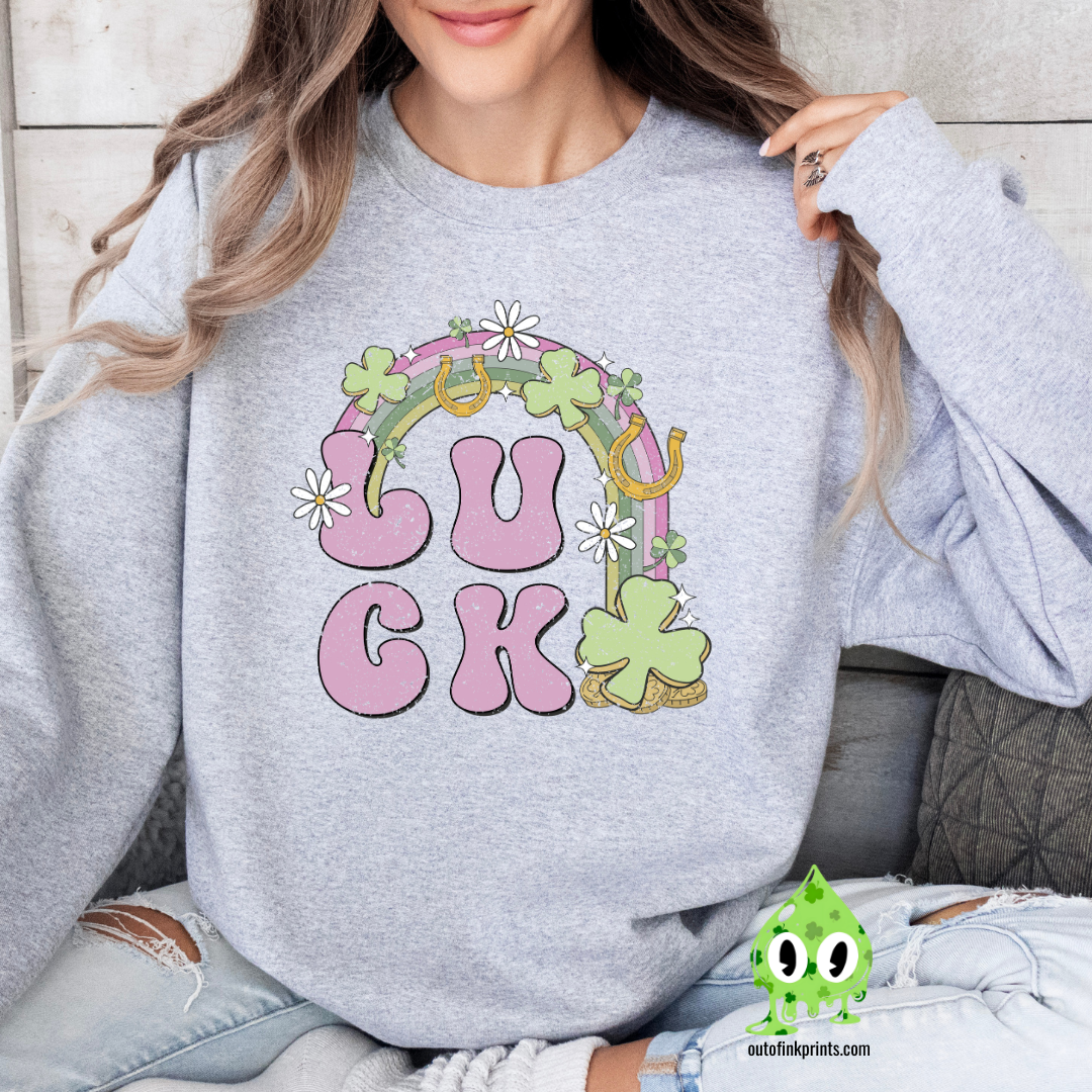 A woman wearing a grey sweatshirt with the text 'LUCK' arranged in a playful design using Irish-themed graphics such as shamrocks and a four-leaf clover.