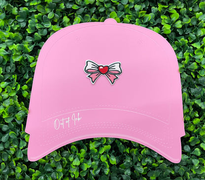A pink hat that has a patch with a pink and white bowtie design and a heart in the center