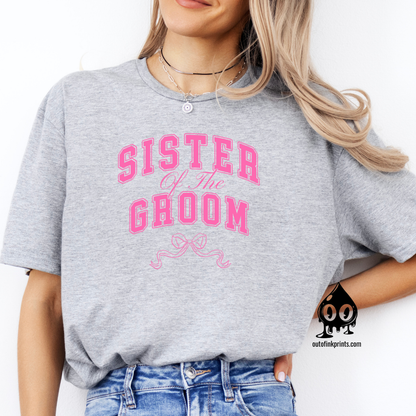 A woman sitting wearing a gray t-shirt with the text 'Sister of the Groom' and a pink bow design on the front.
