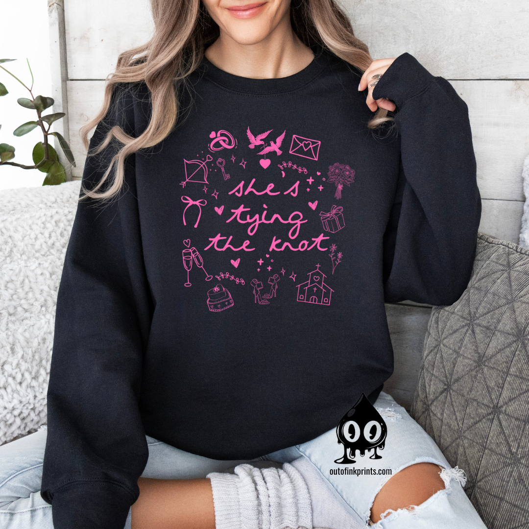 A black text sweatshirt with 'She's Tying the Knot' printed on the front, surrounded by various symbols and illustrations related to weddings.