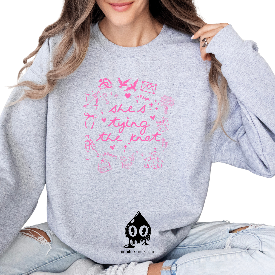 A gray sweatshirt with pink text 'She's Tying the Knot' printed on the front, surrounded by various symbols and illustrations related to weddings.