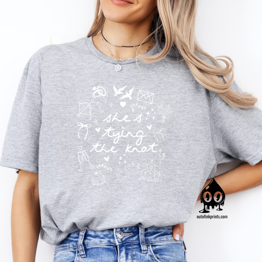 A woman sitting and wearing a gray T-shirt with white text and graphics related to a bride tying the knot.
