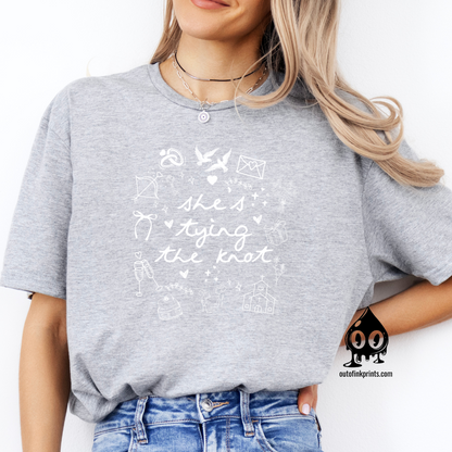 A woman sitting and wearing a gray T-shirt with white text and graphics related to a bride tying the knot.
