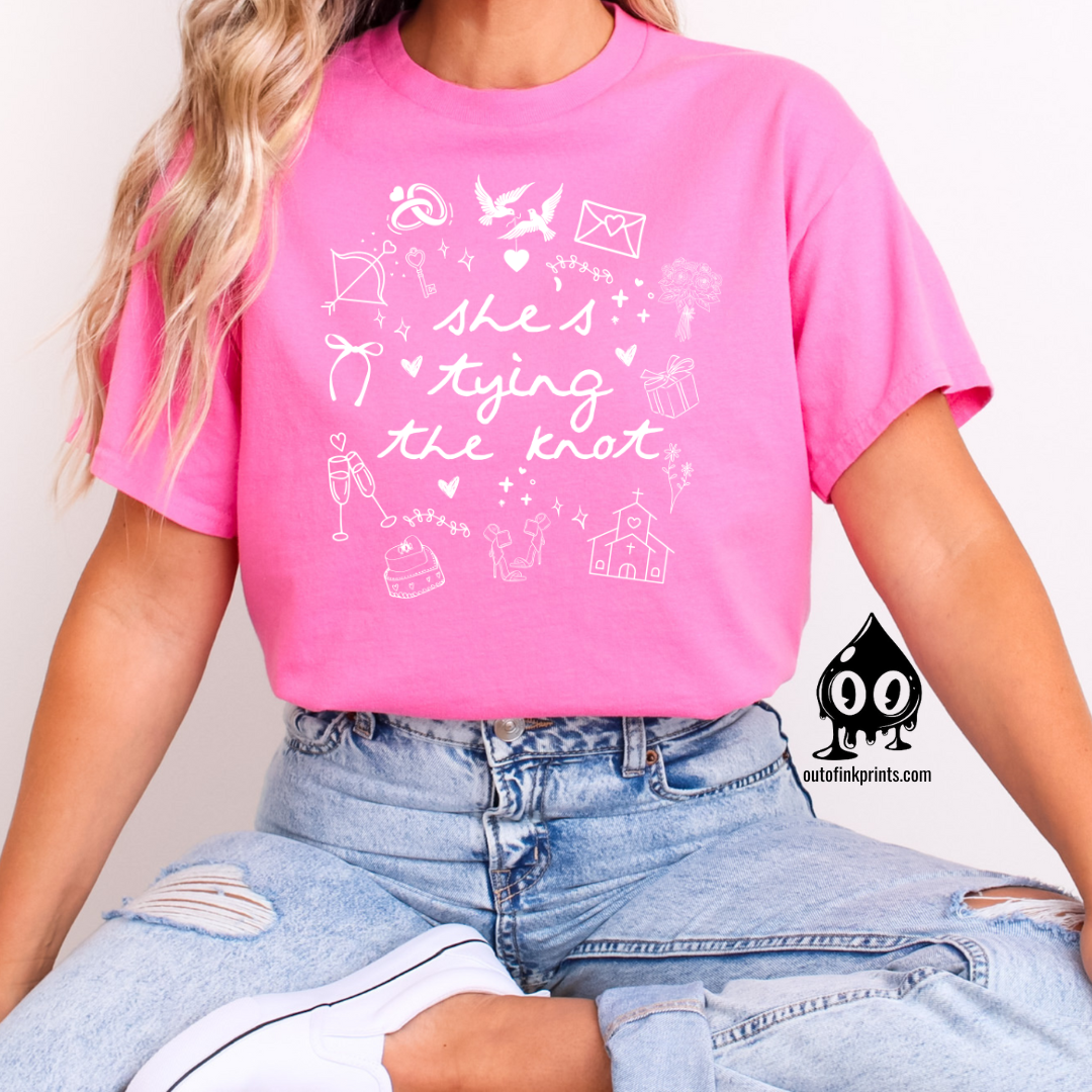 A woman sitting and wearing a pink T-shirt with white text and graphics related to a bride tying the knot.