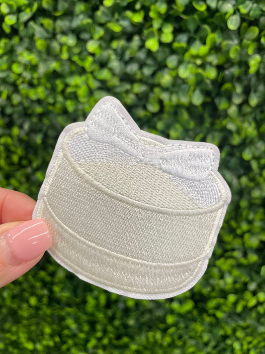 Embroidered patch of a white cake with a white bow placed on top.
