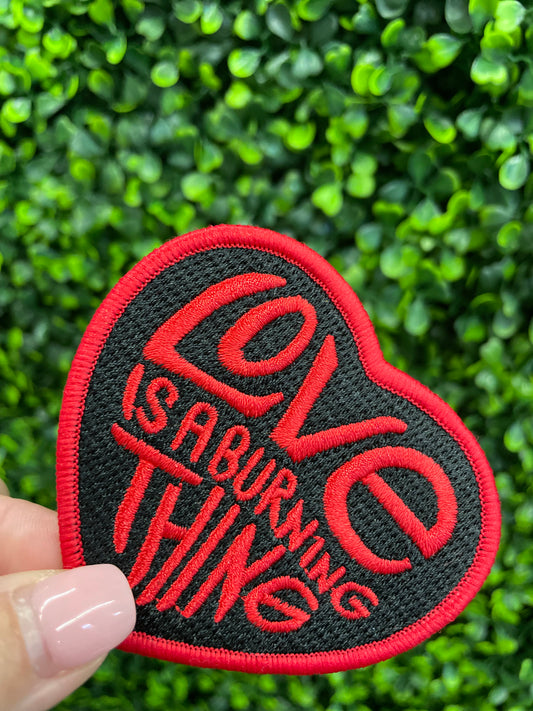 Heart-shaped patch with 'Love is a Burning Thing' text held against a green leafy background