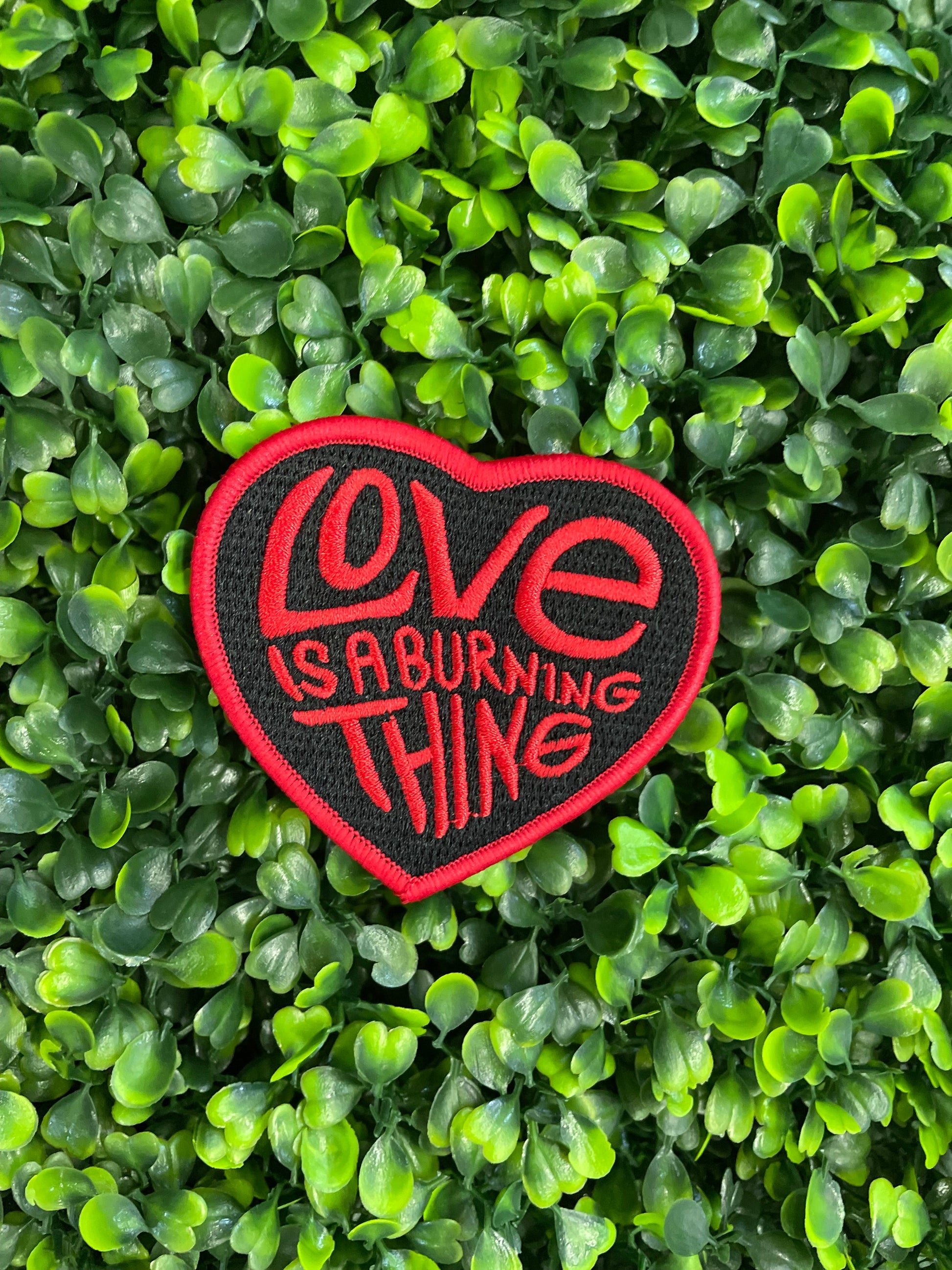 Heart-shaped patch with 'Love is a Burning Thing' text, placed against a green and leafy background