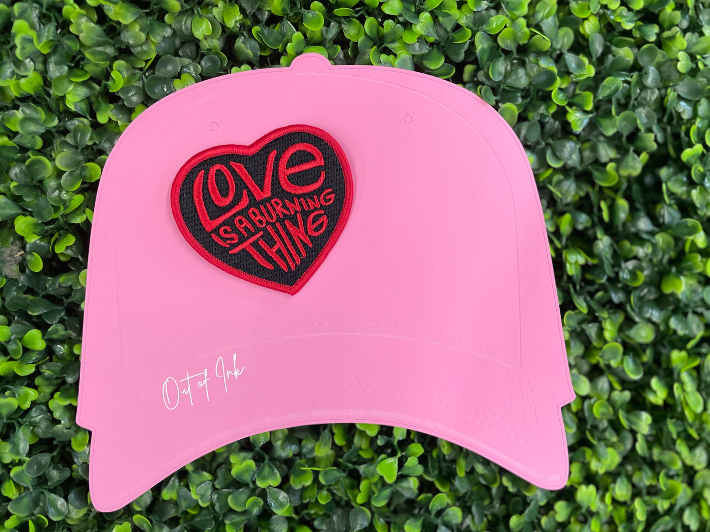 Heart-shaped patch with 'Love is a Burning Thing' text, placed on a pink trucker hat graphic