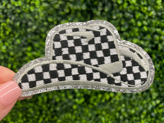 Checkered white and black cowboy hat patch with silver lining, held against a green leafy background