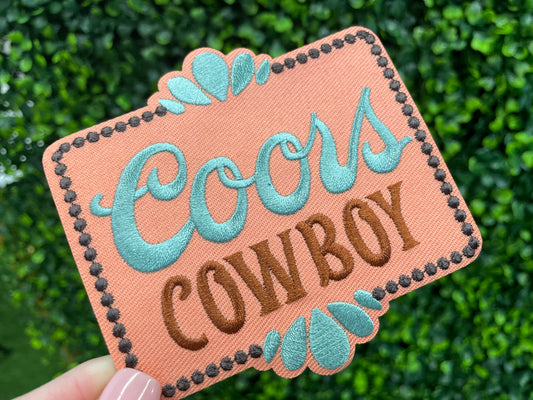 Embroidered patch with 'Coors Cowboy' text on a light orange background with teal accents, held against a green leafy background