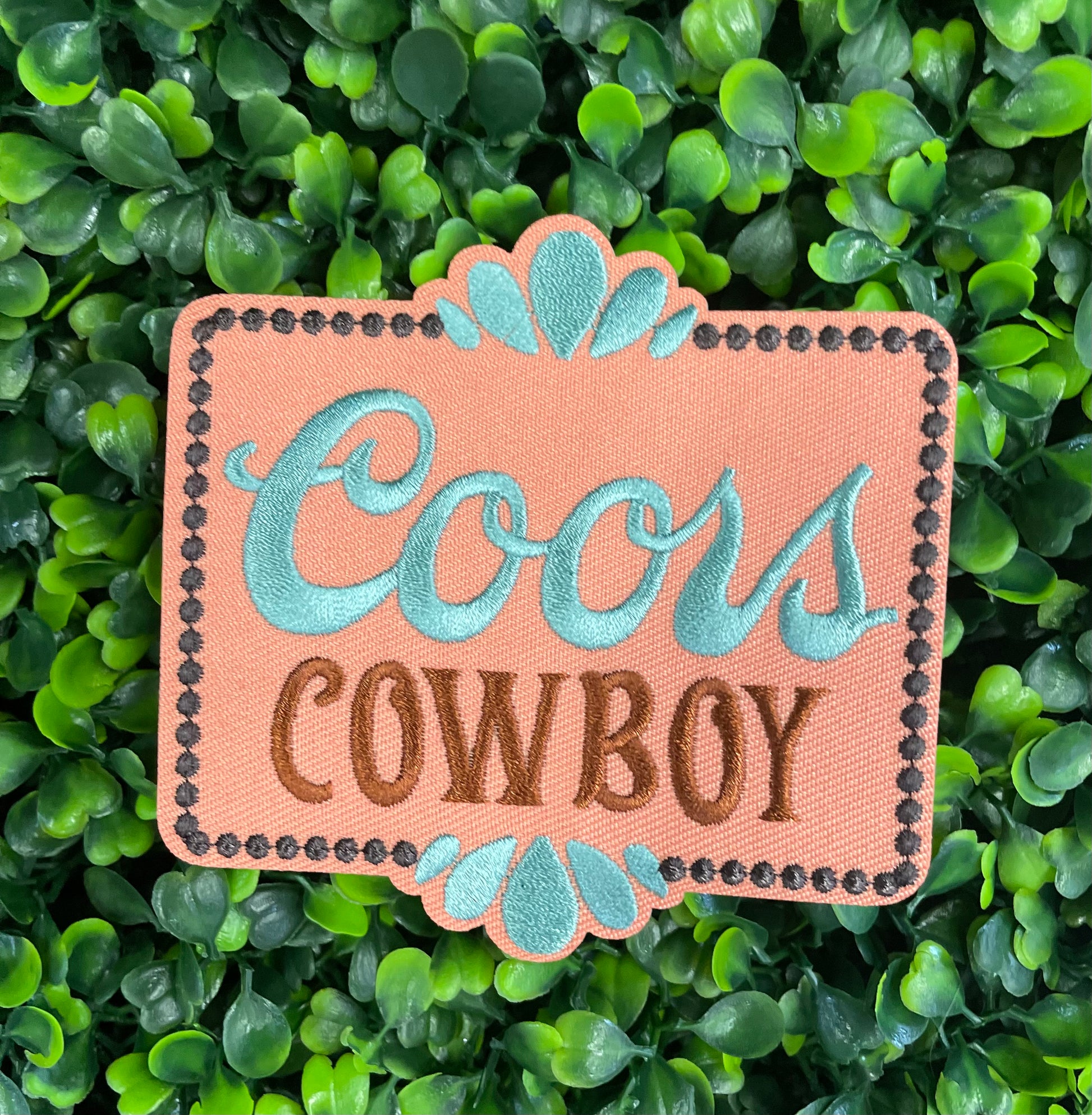 Embroidered patch with 'Coors Cowboy' text on a light orange background with teal accents, placed on a green leafy backdrop