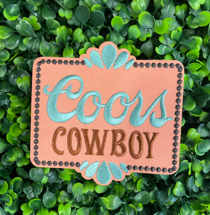 Embroidered patch with 'Coors Cowboy' text on a light orange background with teal accents, placed on a green leafy backdrop