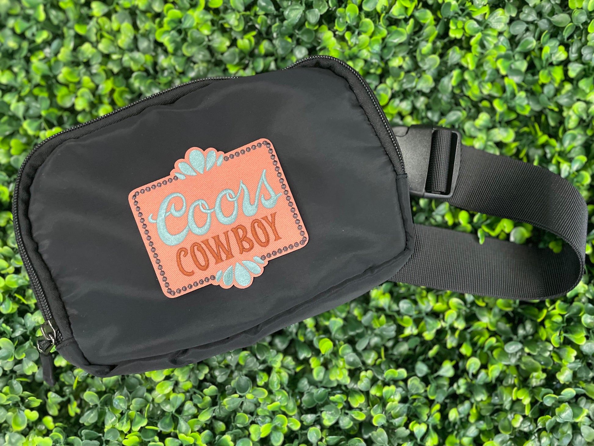 Embroidered patch with 'Coors Cowboy' text on a light orange background with teal accents, placed on a black fanny pack
