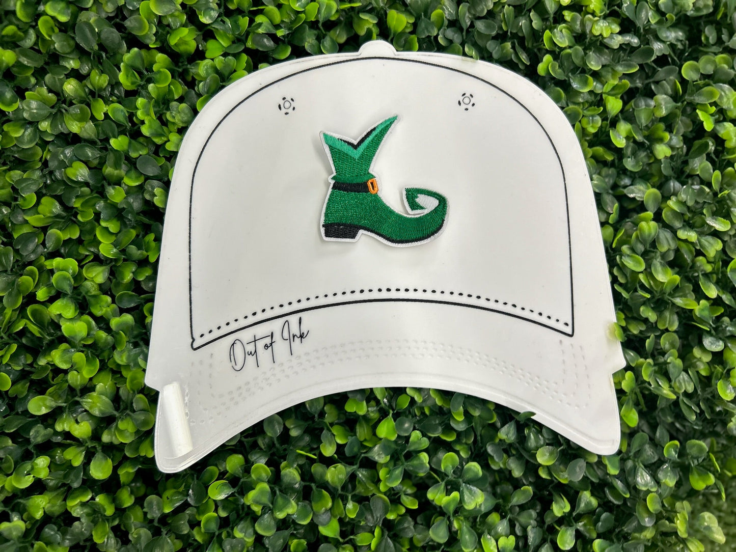 Green elf shoe embroidered patch with curled toe, buckle, and white border held on a white hat graphic