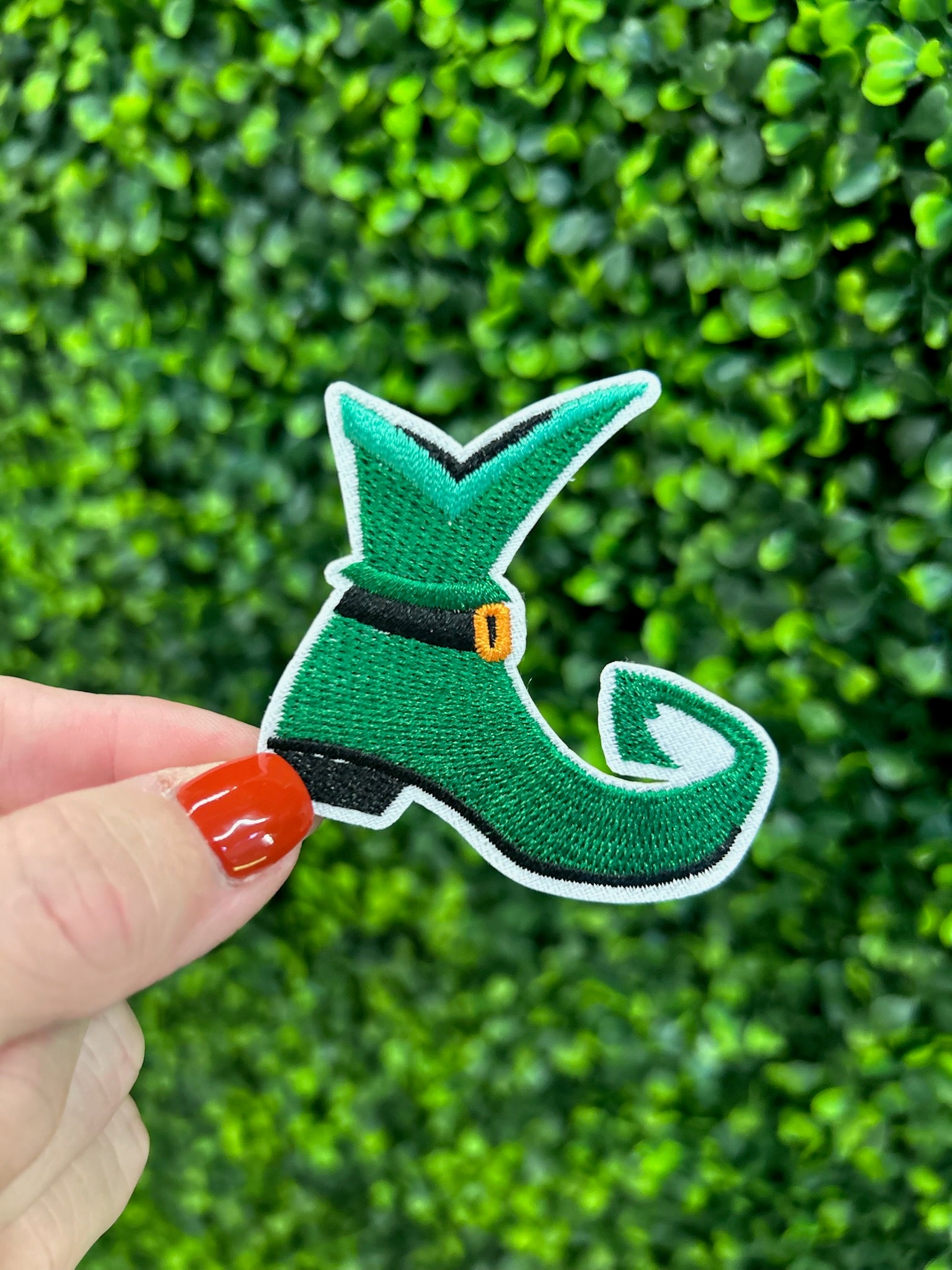 Green elf shoe embroidered patch with curled toe, buckle, and white border. Held against a green leafy background
