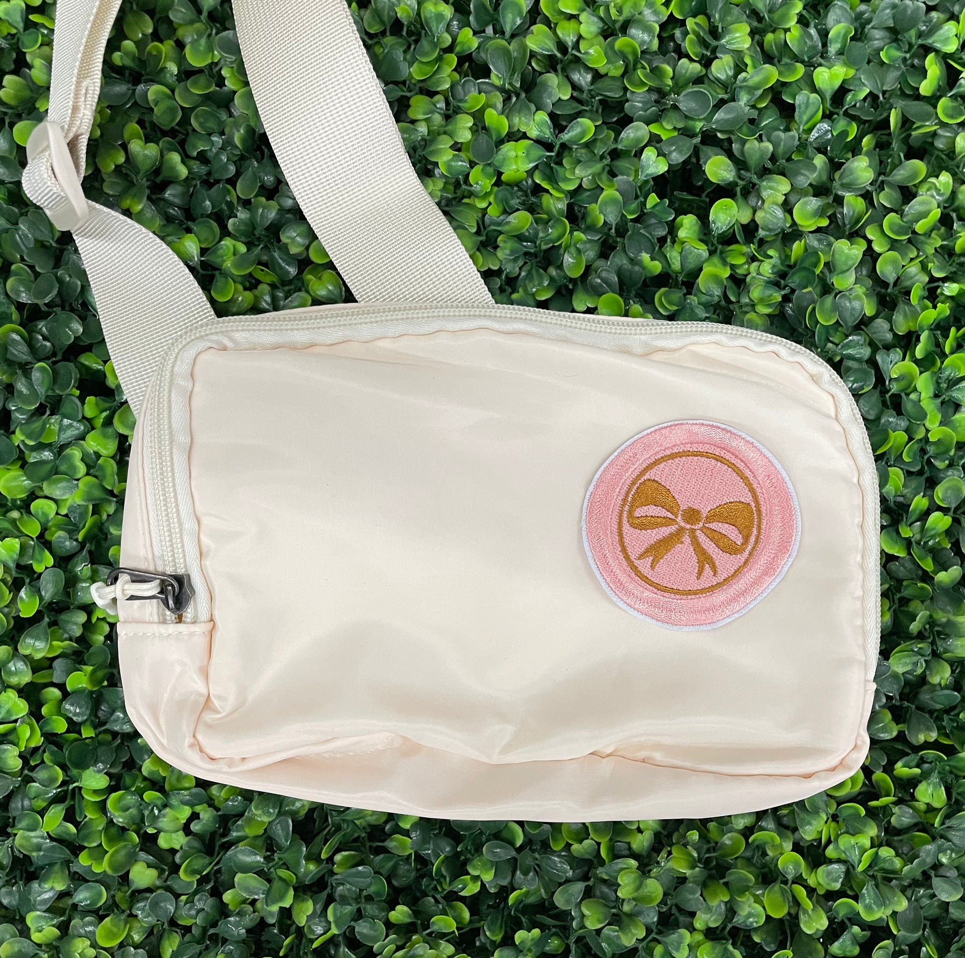 A round patch featuring a gold bow on a pink background with a embroidered edge, placed on a white shoulder bag