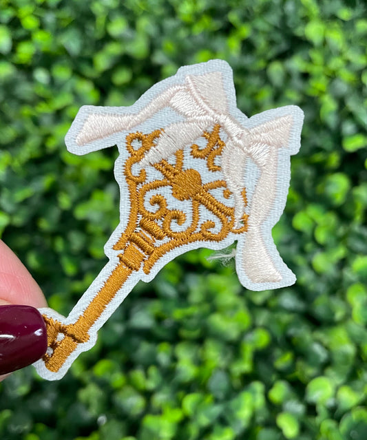 A patch in the shape of a vintage key with a gold bow, embroidery work, on a white background.