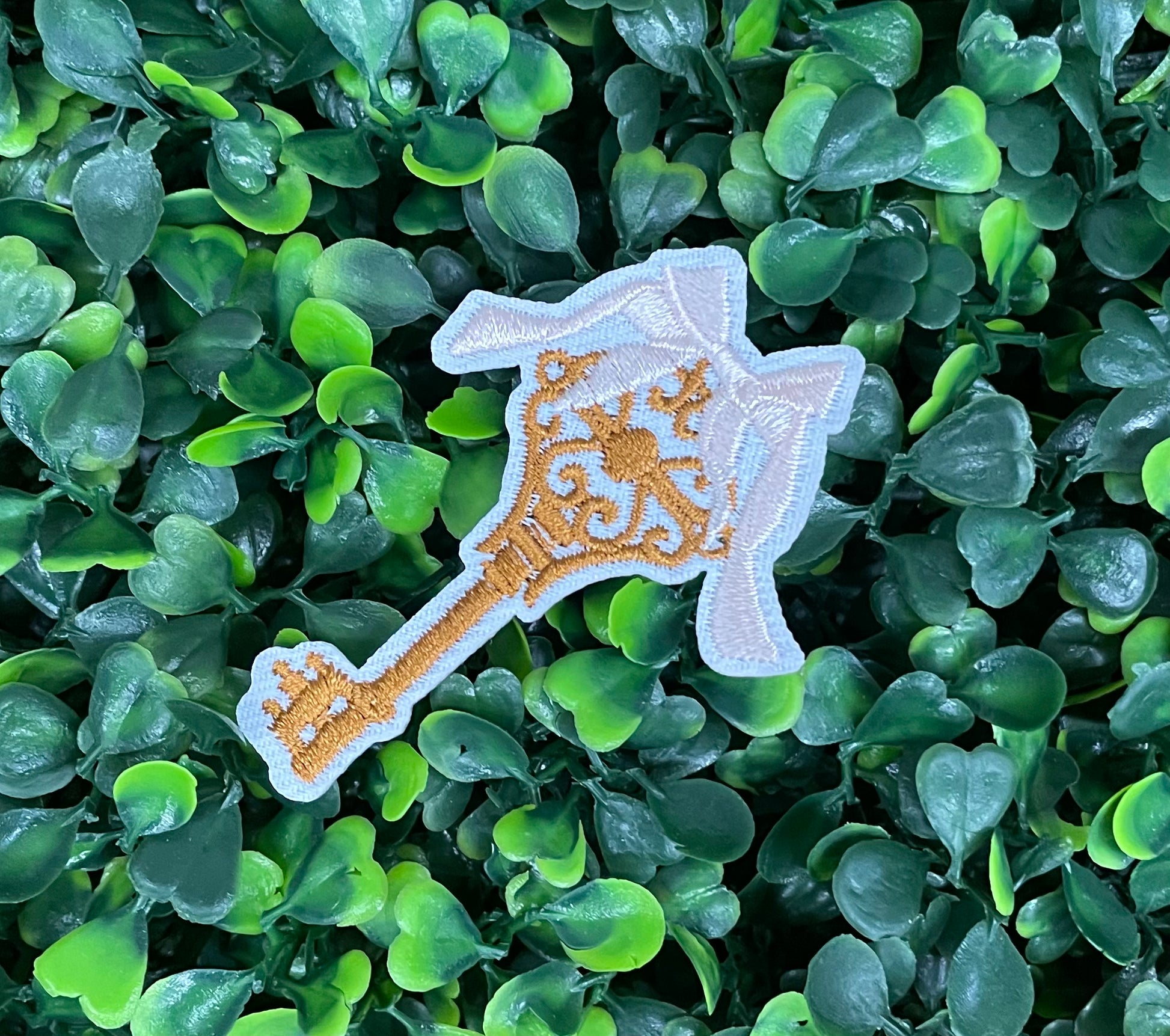 A patch in the shape of a vintage key with a gold bow, embroidery work, set on a background of greenery