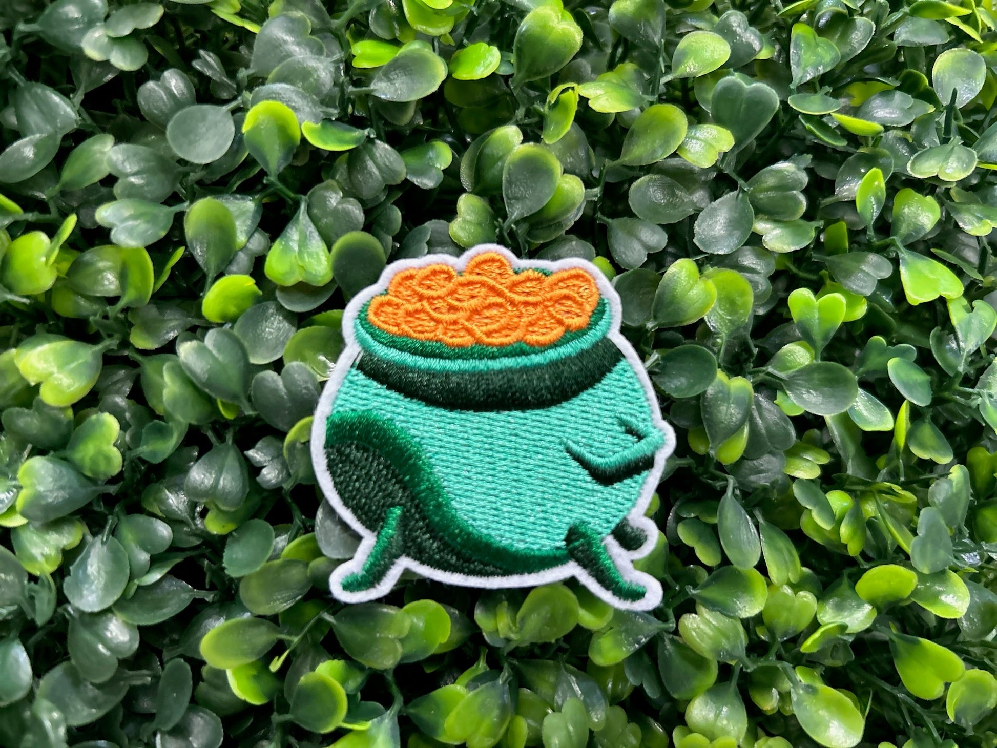 Green cauldron embroidered patch filled with gold coins and white border held on a green and leafy background