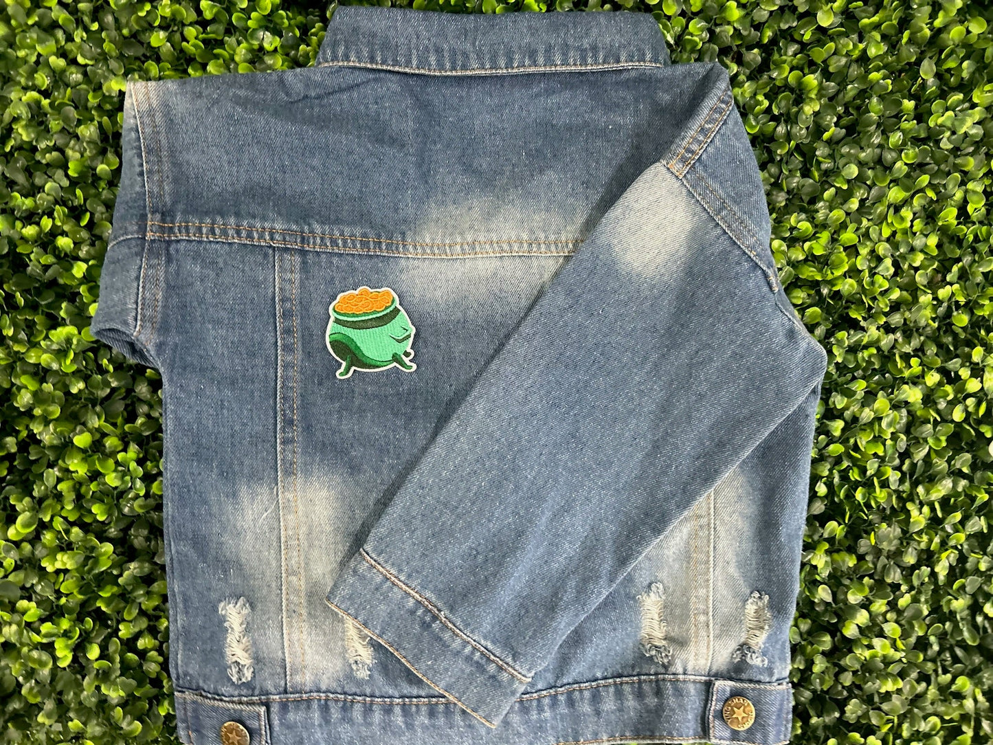 Green cauldron embroidered patch filled with gold coins and white border placed on a children's denim jean jacket