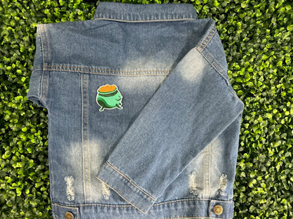 Green cauldron embroidered patch filled with gold coins and white border placed on a children's denim jean jacket