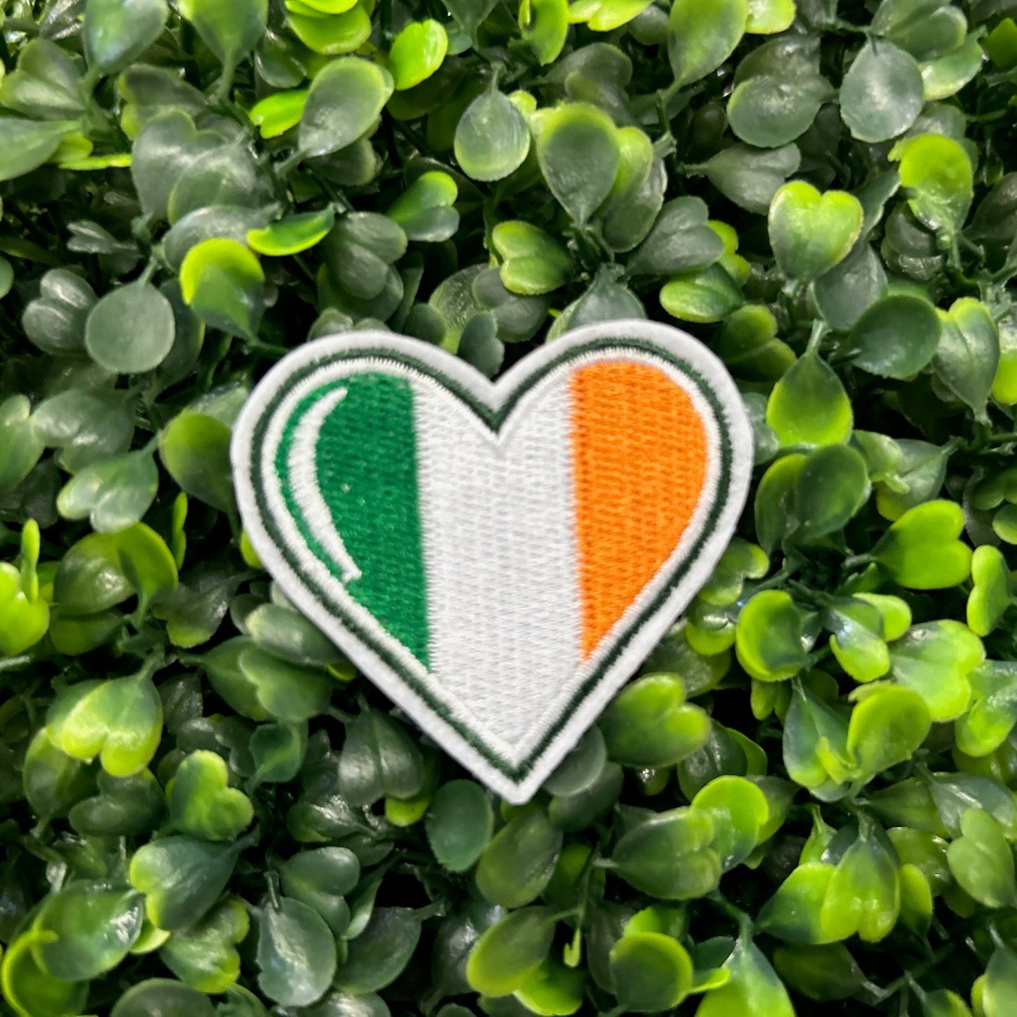 Heart shaped embroidered patch with Ireland flag colors green white and orange held on a green leafy background
