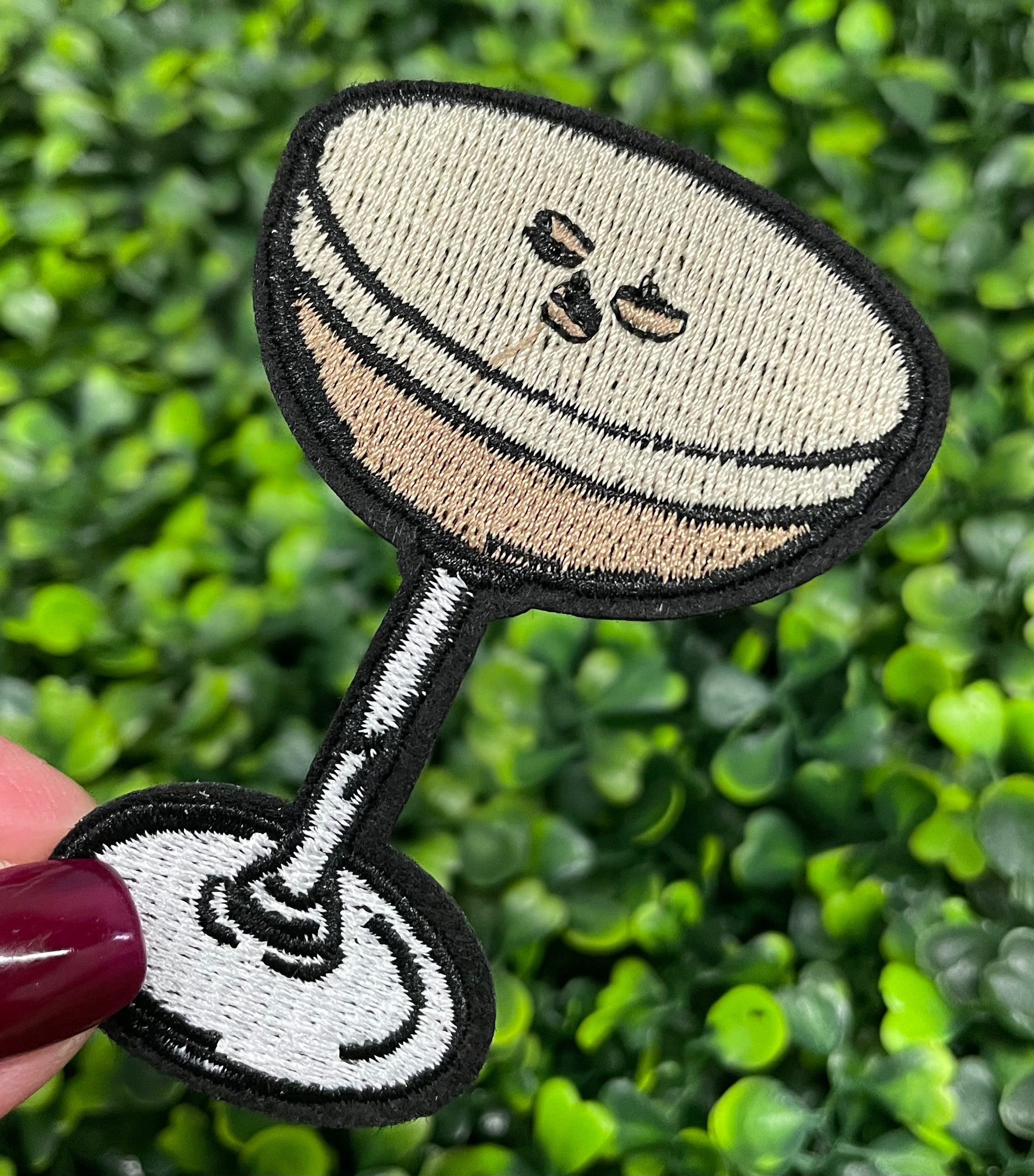 An iron-on patch depicting an espresso martini with a glass and coffee beans, held against a green leafy background.