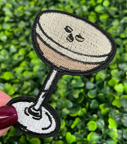 An iron-on patch depicting an espresso martini with a glass and coffee beans, held against a green leafy background.