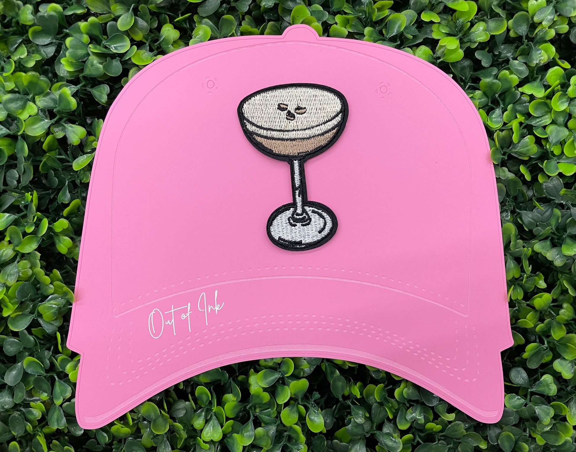 An iron on embroidered martini glass depiction set on a pink hat against a green backdrop