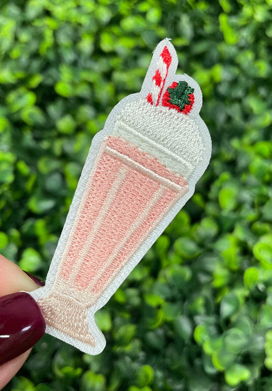 A pink milkshake embroidered patch with a cherry on top, held against a green leafy background.