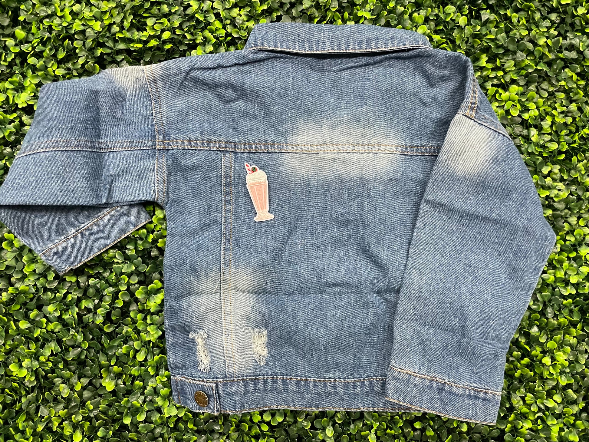 A pink milkshake embroidered patch with a cherry on top, held onto the back of a jean jacket against a green leafy background