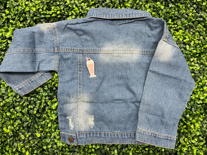 A pink milkshake embroidered patch with a cherry on top, held onto the back of a jean jacket against a green leafy background