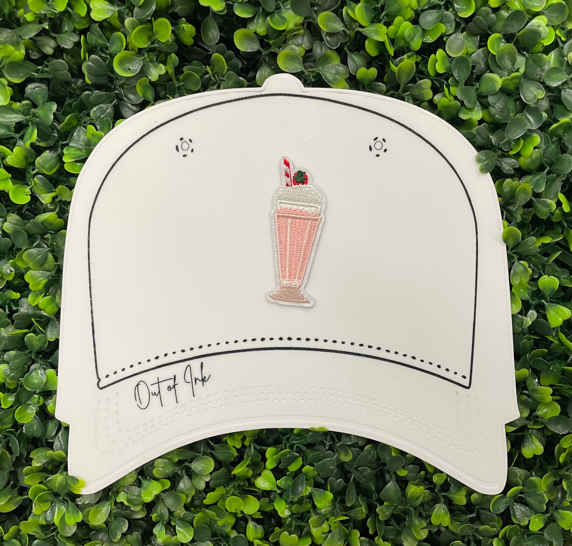 A pink milkshake embroidered patch with a cherry on top, held onto a white hat design 