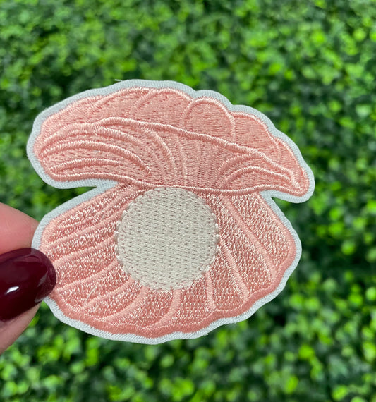 A pink oyster with pearl patch held against a green leafy background, showcasing an embroidered design resembling an oyster shell with a pearl interior.