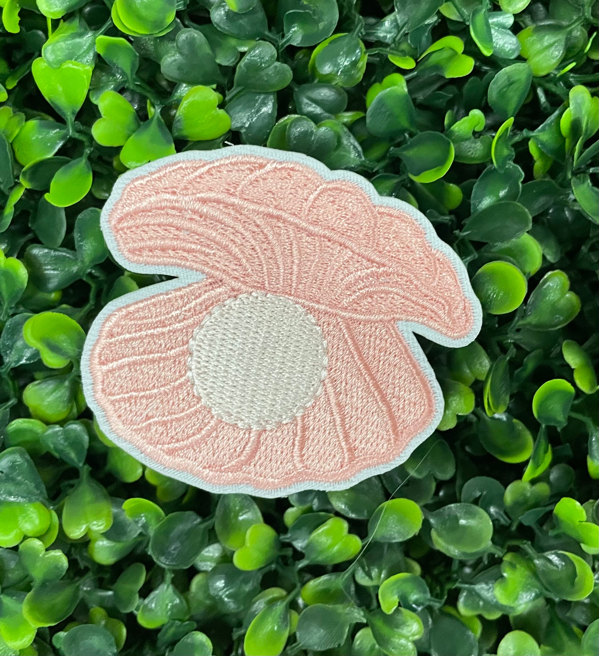 A pink oyster with pearl patch held against a green leafy background, showcasing an embroidered design resembling an oyster shell with a pearl interior.