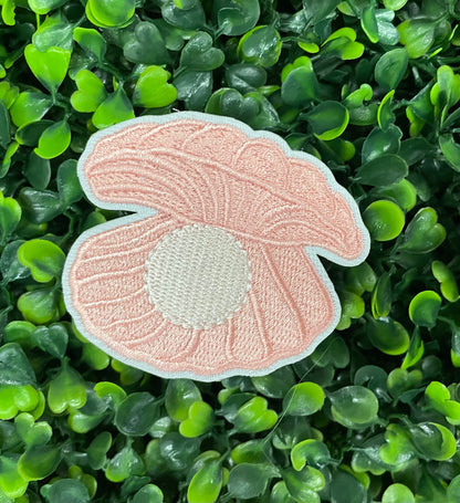 A pink oyster with pearl patch held against a green leafy background, showcasing an embroidered design resembling an oyster shell with a pearl interior.