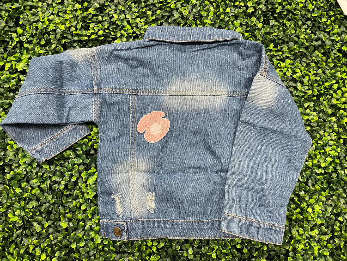A pink patch showcasing an embroidered design resembling an oyster shell with a pearl interior, held onto a jean jacket