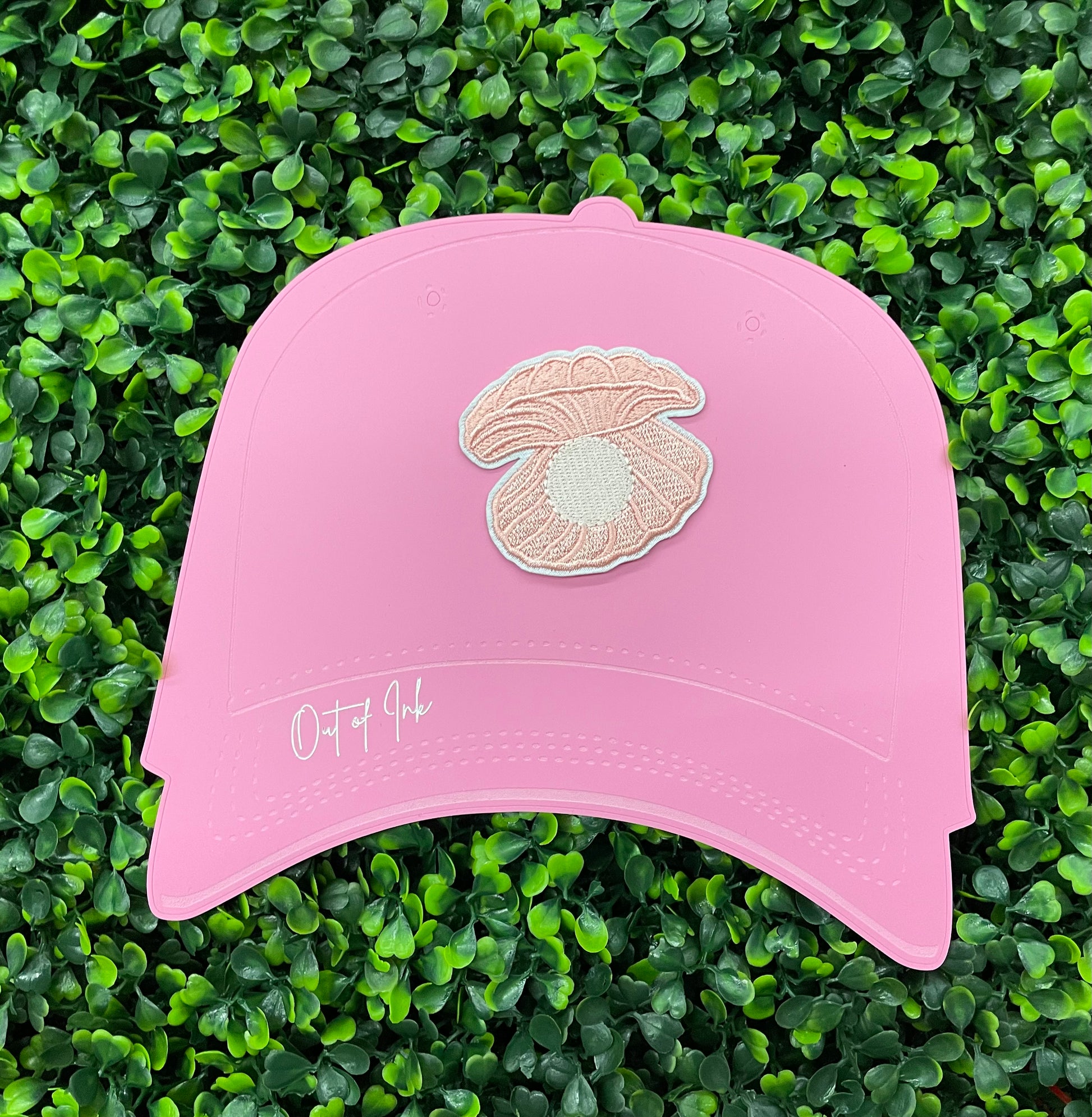 A pink patch showcasing an embroidered design resembling an oyster shell with a pearl interior, held onto a pink hat against a green leafy background