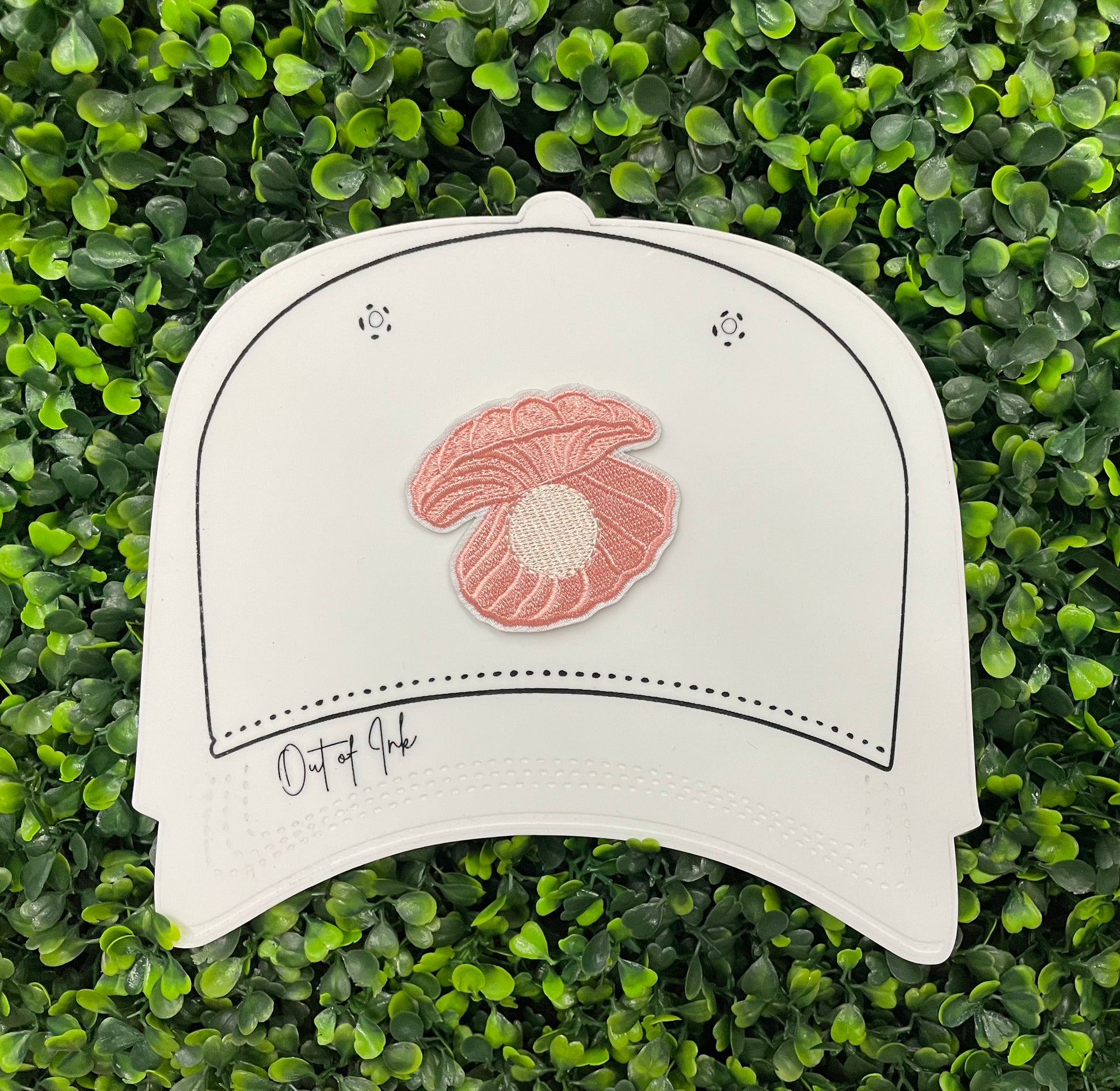 A pink patch showcasing an embroidered design resembling an oyster shell with a pearl interior, held against a white hat