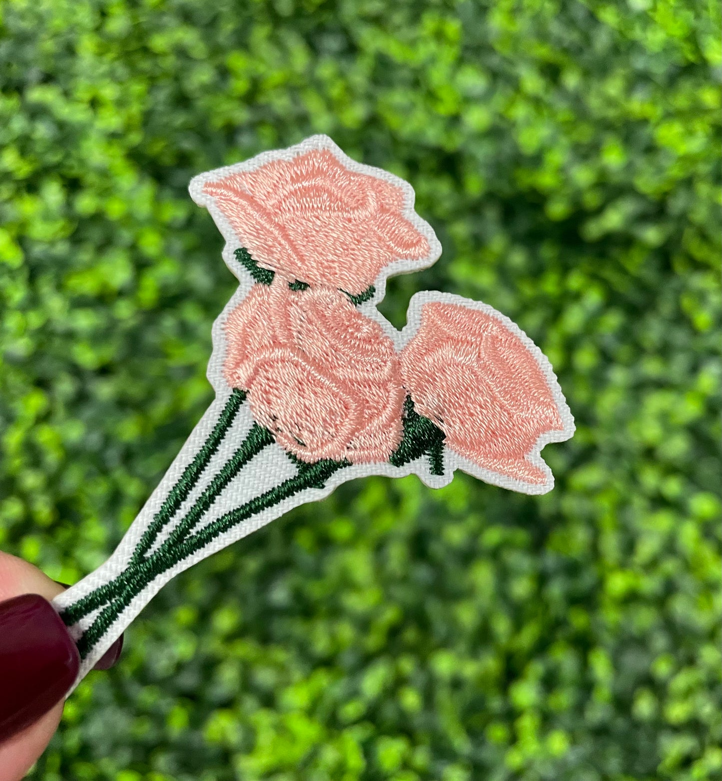 A hand holding an iron-on patch with embroidered pink roses attached to a green stem, set against a background of greenery.