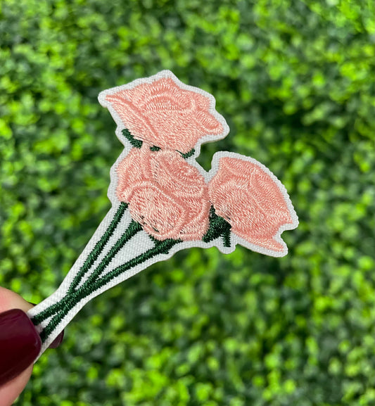 A hand holding an iron-on patch with embroidered pink roses attached to a green stem, set against a background of greenery.