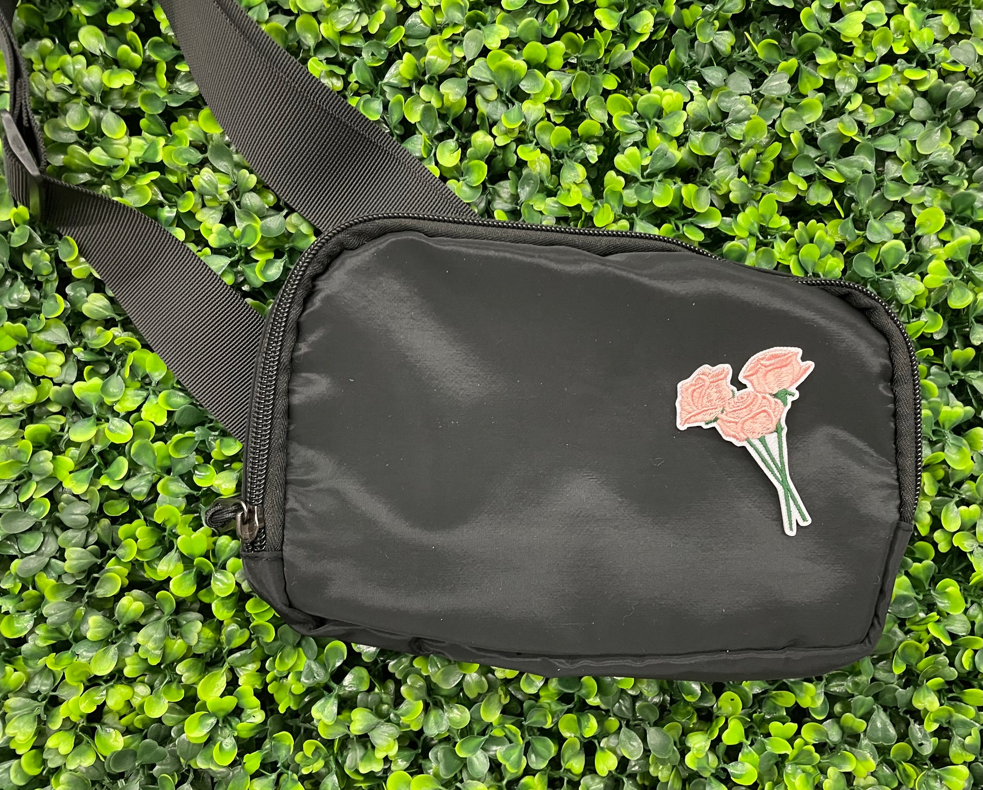 A hand holding an iron-on patch with embroidered pink roses attached to a green stem, set on a black shoulder bag