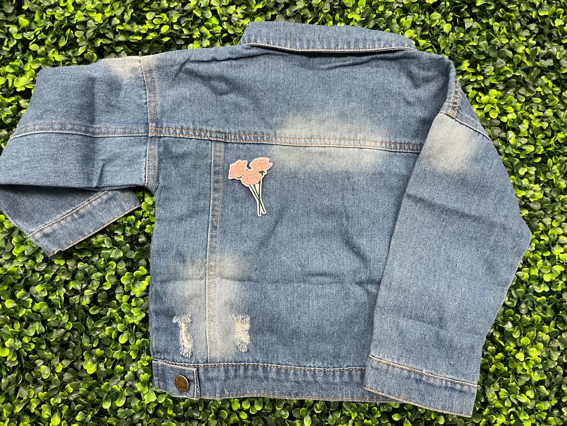 A hand holding an iron-on patch with embroidered pink roses attached to a green stem, set on a jean jacket