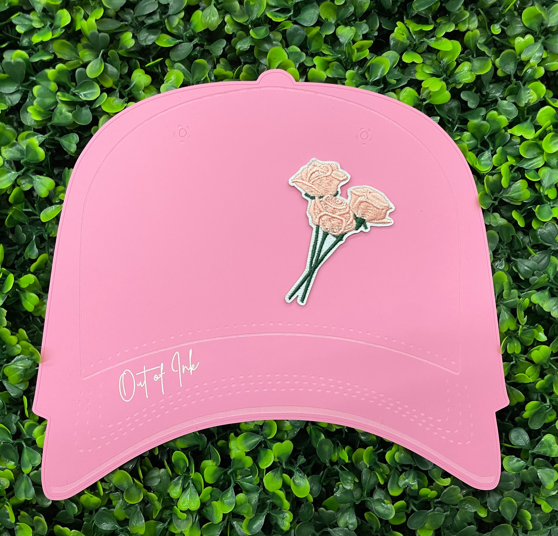 A hand holding an iron-on patch with embroidered pink roses attached to a green stem, set on a pink hat