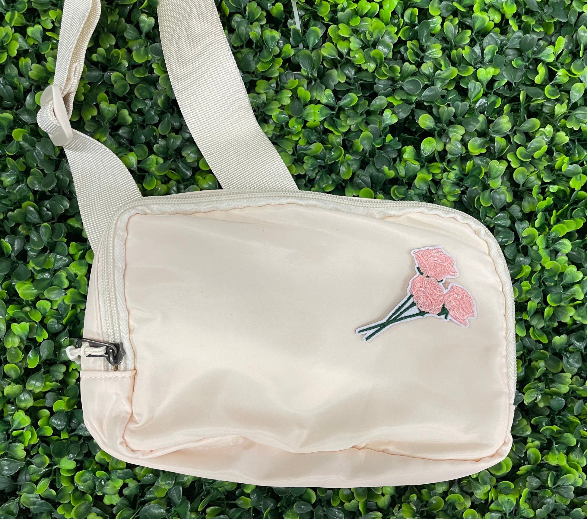 A hand holding an iron-on patch with embroidered pink roses attached to a green stem, set on a white bag