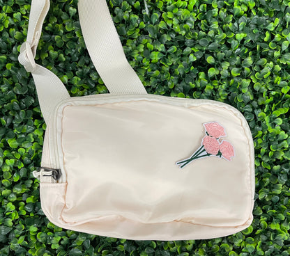 A hand holding an iron-on patch with embroidered pink roses attached to a green stem, set on a white bag