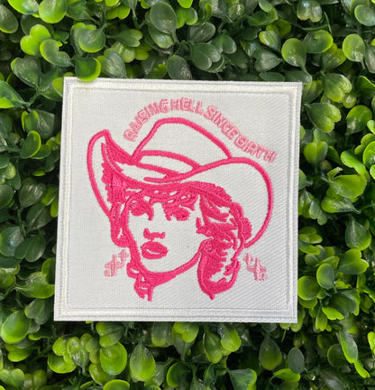 Embroidered patch with pink design and text "raising hell since birth" on a white background, placed on a green leafy backdrop