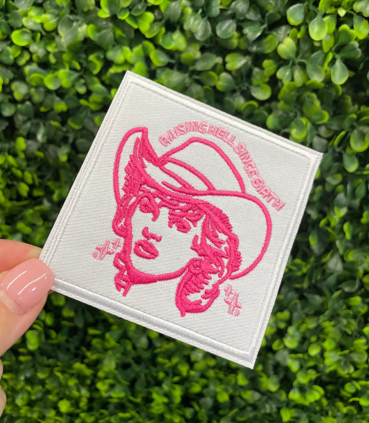 Embroidered patch with pink design and text "raising hell since birth" on a white background, held against a green leafy background.